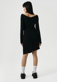 Vrouw staat met haar rug naar ons toe, gekleed in een asymmetrische zwarte jurk met lange mouwen, witte sokken en zwarte chunky schoenen op een effen achtergrond.
