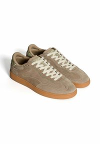Un paio di sneaker in suede beige con lacci bianchi e suole in gomma, esibite su uno sfondo bianco.
