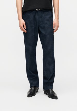 Άνδρας που φοράει σκούρα ριγέ παντελόνια με φαρδιά μπατζάκια και πλακέ τσέπες, μαύρη ζώνη, μαύρο πουκάμισο και μαύρα μυτερά δερμάτινα παπούτσια.