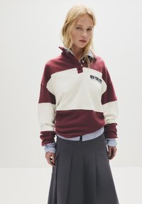 Pullover a blocchi di colore bordeaux e bianco con colletto e testo "New York City"; abbinato a una gonna plissettata scura e polsini a righe.