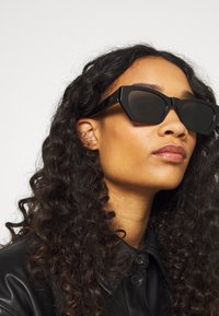 Occhiali da sole neri e angolari con lenti scure, caratterizzati da un design audace. Chi li indossa ha i capelli lunghi e ricci ed è vestito con una giacca di pelle nera.