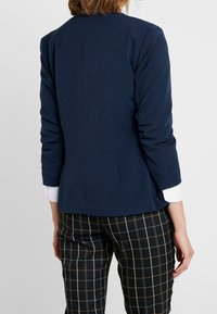 Blazer blu navy con una texture liscia, design aderente e maniche a tre quarti. Abbinato a pantaloni a scacchi bianco e nero con linee sottili.