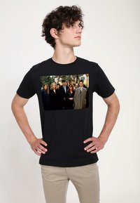 Camiseta negra con un gráfico rectangular de un grupo vestido con ropa oscura, rodeado de vegetación. Tela suave; corte clásico.