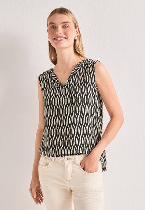 Jeune femme aux longs cheveux blonds portant un haut sans manches noir et blanc à motifs et un pantalon beige clair, souriante avec une main dans la poche.