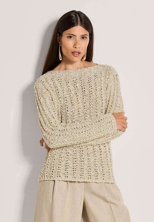 Frau mit langen dunklen Haaren, die einen beigen Strickpullover, beige weite Hosen und große goldene Hängeohrringe trägt, posiert mit verschränkten Armen.
