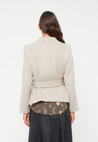Giacca grigia chiara con cintura, spalle strutturate, tessuto testurizzato e vestibilità sartoriale, indossata sopra una blusa scura a fantasia e una gonna nera.