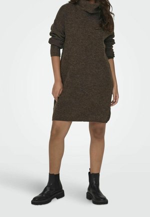 Kvinde iført en brun strikket kjole med opkørte ærmer og sorte ankelsstøvler, stående mod en ensartet lys baggrund.