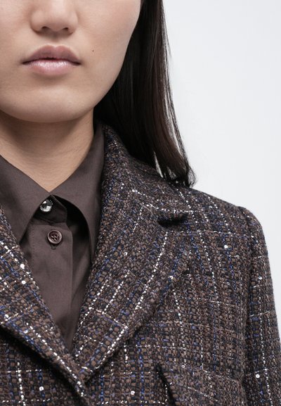 Pruun tweedjakk tekstureeritud kangaga, milles on siniseid ja hõbedasi niite. Märkimisväärse tipu-lapiga ja all on tume nööbitav kraega särk.