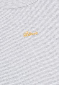 Valge vahvlitekstiil, mille keskel on väikesed kollased kalligraafilised sõnad "Blkvis" brodeeritud.