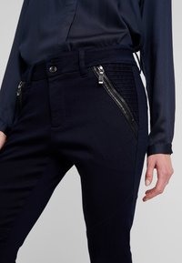 Kvinna iklädd mörkblå byxor med diagonala dragkedjefickor framtill och en matchande långärmad blus, med handen avslappnat vid sidan.