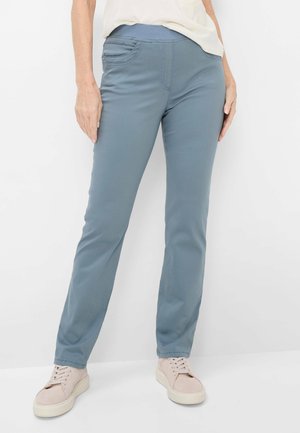 Kvinde iført strækbare blågrå bukser med elastisk taljebånd og lyserøde snøresko, stående foran en ensfarvet hvid baggrund.