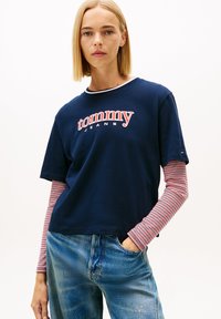 Granatowy t-shirt z krótkim rękawem z czerwono-białym logo "tommy JEANS". Nałożony na czerwono-białe paskowane długie rękawy, w zestawieniu z niebieskimi dżinsami.
