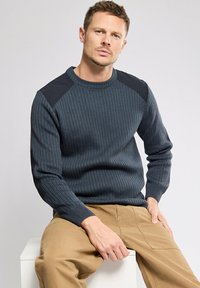 Dunkelgrauer, gerippter Pullover mit schwarzen Schulterakzenten, Rundhalsausschnitt und langen Ärmeln, kombiniert mit khakifarbenen Hosen. Strukturiertes Material.