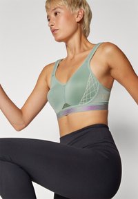 Sujeción deportiva de color verde claro con tela texturizada, diseño de escote en V y patrón geométrico. Presenta una banda inferior acentuada en púrpura. Leggings negros.