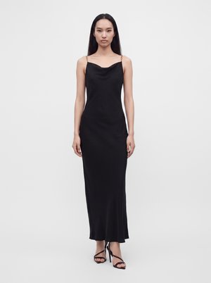 Femme debout, face à l'objectif, portant une longue robe noire à fines bretelles en chaîne et des sandales noires à talons hauts et à lanières.