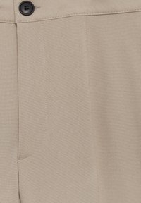 Detailní záběr na béžovou látku ukazující obrys prošité kapsy a černý knoflík s čtyřmi otvory na pásku u klasických kalhot.