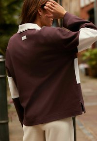 Dunkelbrauner Pullover mit weißen Akzenten, ausgestattet mit einem klassischen Kragen, lockerer Passform und seitlichen Schlitzen. Der Stoff wirkt weich und strukturiert.