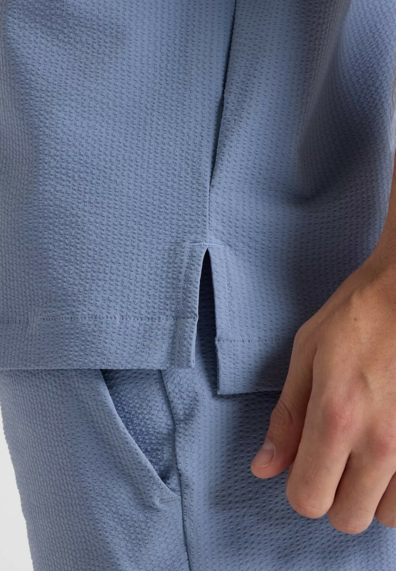 Chemise bleu clair texturée avec une fente latérale et une poche visible. Le tissu présente un motif gaufré subtil et une finition lisse.
