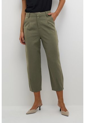 Femme portant un pantalon court vert olive sur mesure avec des plis à l'avant et des talons slingback beiges pointus, debout sur un fond blanc.