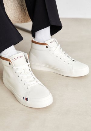 Białe sneakersy typu high-top z brązowymi wstawkami, białymi sznurówkami i logo „Hilfiger”, noszone z czarnymi spodniami i białymi skarpetkami.