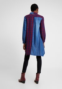 Persona che indossa un lungo vestito camicia patchwork con pannelli scozzesi rossi e denim blu, pantaloni neri e stivaletti rossi, di spalle.