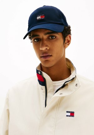 Cappellino da baseball navy con logo "TOMMY JEANS"; giacca beige con colletto alto, chiusura con zip e accenti colorati in rosso e navy.