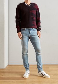 Bordový a černý pletený svetr s v-výstřihem a bílým lemem, spárovaný s světle modrými slim jeans a bílými teniskami s hnědými akcenty.