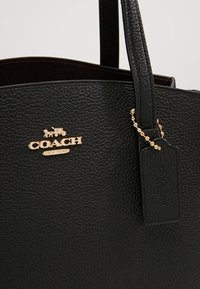 Sac cabas Coach en cuir texturé noir avec logo doré et emblème de calèche, doté d'une étiquette en cuir attachée par une chaîne perlée dorée.