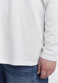 Weißes langärmliges Hemd mit Rundhalsausschnitt aus weichem Baumwollstoff. Ein kleines Logo-Patch ist am Saum sichtbar. Getragen mit blauen Jeans.