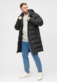 Cappotto puffo nero con cappuccio, caratterizzato da un design trapuntato e chiusura frontale con zip. Indossato sopra un felpa grigia e jeans blu, abbinato a sneakers bianche.