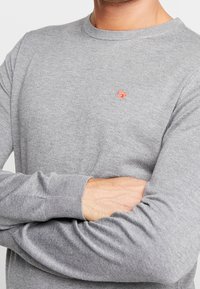 Grauer gestrickter Pullover mit rundem Halsausschnitt, gerippten Bündchen und einem kleinen orangefarbenen Logo auf der Brust. Der Stoff wirkt weich und leicht.