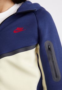 Marineblå hoodie med kremfarvet panel, rød Nike-logo og grå lynlås-lomme på ærmet. Glat stof med kontrasterende farveblokdesign.