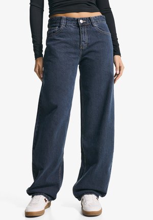 Jeans a gamba larga di colore blu scuro in denim, con chiusura a bottone e cuciture a contrasto. Abbinati a sneakers bianche.