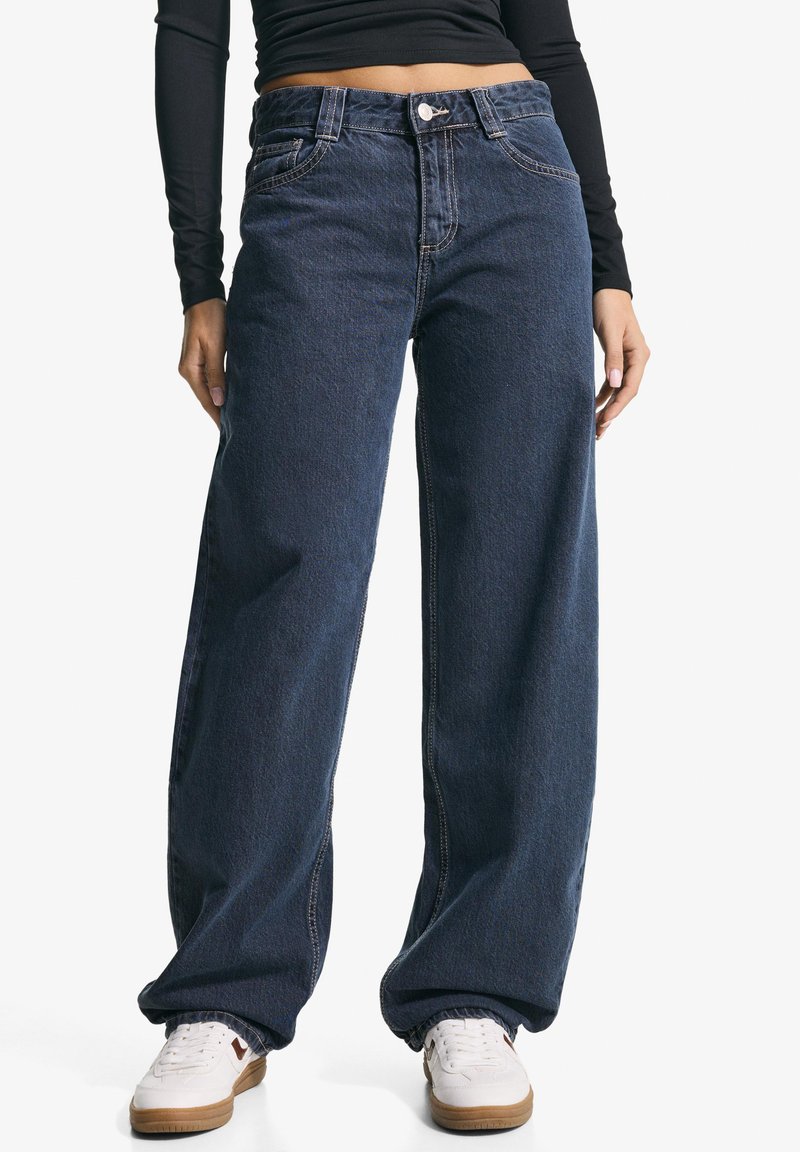 Široké tmavě modré džíny vyrobené z denimu, s knoflíkovým zapínáním a kontrastním šitím. Vytvořeno v kombinaci s bílými teniskami.