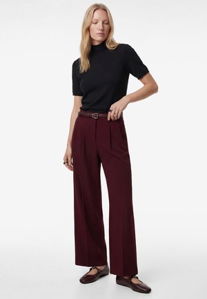 Femme portant un col roulé noir à manches courtes, un pantalon large rouge foncé taille haute avec une ceinture, et des chaussures plates rouges foncées.