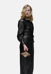 Robe en cuir noir avec une taille ceinturée, des manches longues et une fermeture à boutons. Le mannequin tient une pochette transparente avec une surface réfléchissante.