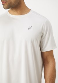 Biały t-shirt sportowy z okrągłym dekoltem, krótkimi rękawami i delikatną teksturą. Posiada małe szare logo na lewej piersi.
