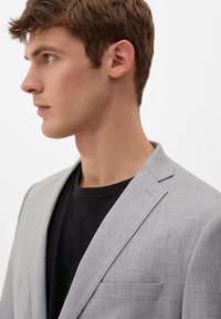 Hellgrauer strukturierter Blazer mit Reverskragen, getragen über einem schwarzen T-Shirt. Verfügt über eine einzelne Brusttasche und eine taillierte Passform.