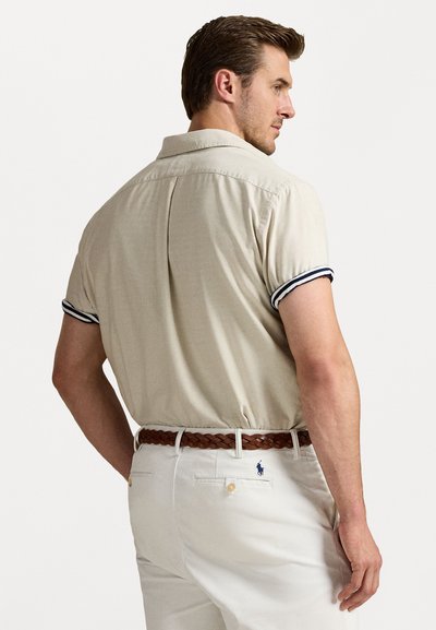 Beige short-sleeve polo with navy-striped cuffs, featuring a back pleat and a brown braided belt over white trousers with a logo detail.