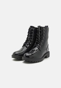 Botas de combate de charol negro con diseño de cordones, ojales de metal y suela gruesa texturizada. Presenta costuras decorativas y una lengüeta.