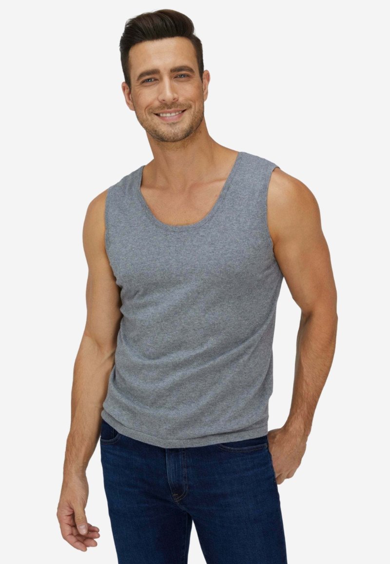 Uomo sorridente con capelli corti e scuri che indossa una canotta grigia e jeans blu scuro, in piedi con una mano in tasca.