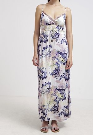 Femme portant une robe sans manches longue jusqu'au sol, à motifs floraux violets et pastel, avec de fines bretelles, assortie à des sandales ouvertes.