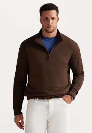 Bruine zip-neck pullover van een textuurstof, gecombineerd met een blauwe T-shirt eronder. Model draagt witte broeken en een horloge.