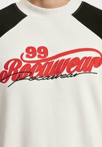 Hvit hettegenser med svarte raglan-ermer. Har rød brodert tekst "99 Bocaweare" i fet retro skrift på brystet.