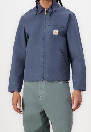 Homme portant une veste bleue zippée avec le logo Carhartt sur la poche poitrine et un pantalon vert sur un fond uni.