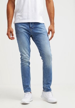 Man die lichtblauwe slim-fit jeans, witte sneakers en een wit T-shirt draagt, staand tegen een effen achtergrond.