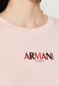 Zbliżenie na osobę w jasnoróżowej koszuli z haftowanym logo "ARMANI EXCHANGE" w czerwonym i czarnym kolorze na piersi.