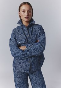Denim jacket featuring a floral pattern in varying shades of blue, with a high collar, front pockets, and metallic button accents.