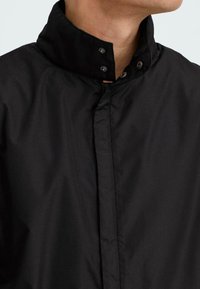 Black waterproof jacket featuring a high collar, snap buttons, and a sleek fabric texture. Designed for functional wear.