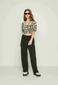 Top curto preto com flores e mangas bufantes, combinado com calças largas pretas e sandálias brancas robustas. Modelo usa óculos de sol escuros.